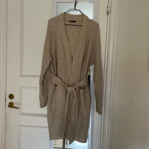 Stickad kofta strl M - En stickad kofta i lång modell från Ginatricot i storlek M. Mjuk och skön med band att knyta i midjan samt fickor fram. I gott skick. 