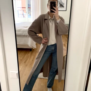 Lång vårig kappa - En längre gråbeige kappa. Har nedhasade axelsömmar som får den att upplevas inte så ”strikt”, sitter så snyggt både om man väljer att ha den öppen eller knyta skärpet antingen baktill eller fram. Har en slits bak. 