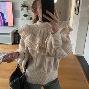 Beige stickad tröja med volangdetaljer - Säljer en superfin beige stickad tröja med volangdetaljer över bröstet och axlarna. Tröjan har långa ärmar och en ribbad struktur som ger en mysig känsla. Super snygg till både vardags och till finare tillställningar! Nästan aldrig använd så är i superfint skick!!💕