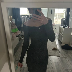 Glittrig svart klänning - Snygg glittrig svart klänning med långärmad design och en tight passform. Perfekt för en kväll ute med en elegant touch. Klänningen har en offshoulder-stil som ger en chic look.