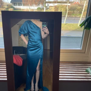 Balklänning  - Elegant blå långklänning från Boohoo med en slits och en one-shoulder design. Klänningen har en glansig finish och är perfekt för speciella tillfällen. Den har en puffärm som ger en unik touch.