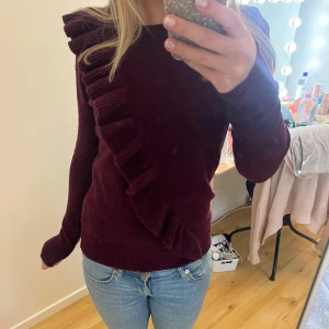 Snygg vinröd tröja med volangdetalj - Snygg vinröd tröja med unik volangdetalj som går diagonalt över framsidan. Tröjan har långa ärmar och är perfekt för att ge en stilfull touch till din outfit.