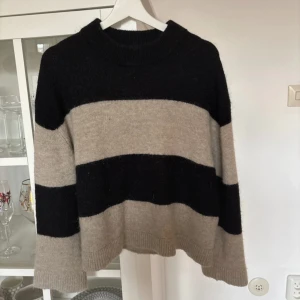 Randig stickad tröja från Gina Tricot - Mysig stickad tröja från Gina Tricot i svart och beige med breda ränder. Tröjan har en avslappnad passform och långa ärmar, perfekt för kyligare dagar. Den är tillverkad i ett mjukt material som ger en skön känsla. Ett måste för garderoben!