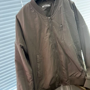 Brun bomberjacka från Gina Tricot - Snygg brun bomberjacka från Gina Tricot med dragkedja framtill och en extra ficka med dragkedja på bröstet. Perfekt för en cool och avslappnad stil. Jackan har långa ärmar och ribbade muddar vid ärmslut och nederkant.