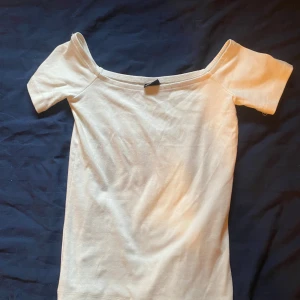 Vit offshoulder topp från Gina Tricot - Säljer en stilren vit offshoulder topp från Gina Tricot.  Inte använd en ända gång. Perfekt för en avslappnad look med korta ärmar och mjukt material. Passar bra till både jeans och kjol.
