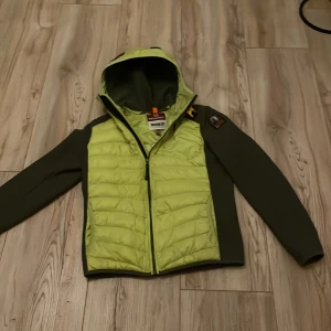 Grön jacka med huva från Parajumpers - Säljer en grön jacka med huva från Parajumpers. Jackan har en cool kombination av ljusgrönt och mörkgrönt med en dragkedja framtill. Märkeslogga på ärmen ger en extra touch. Perfekt för dig som vill ha stil och funktion i ett!