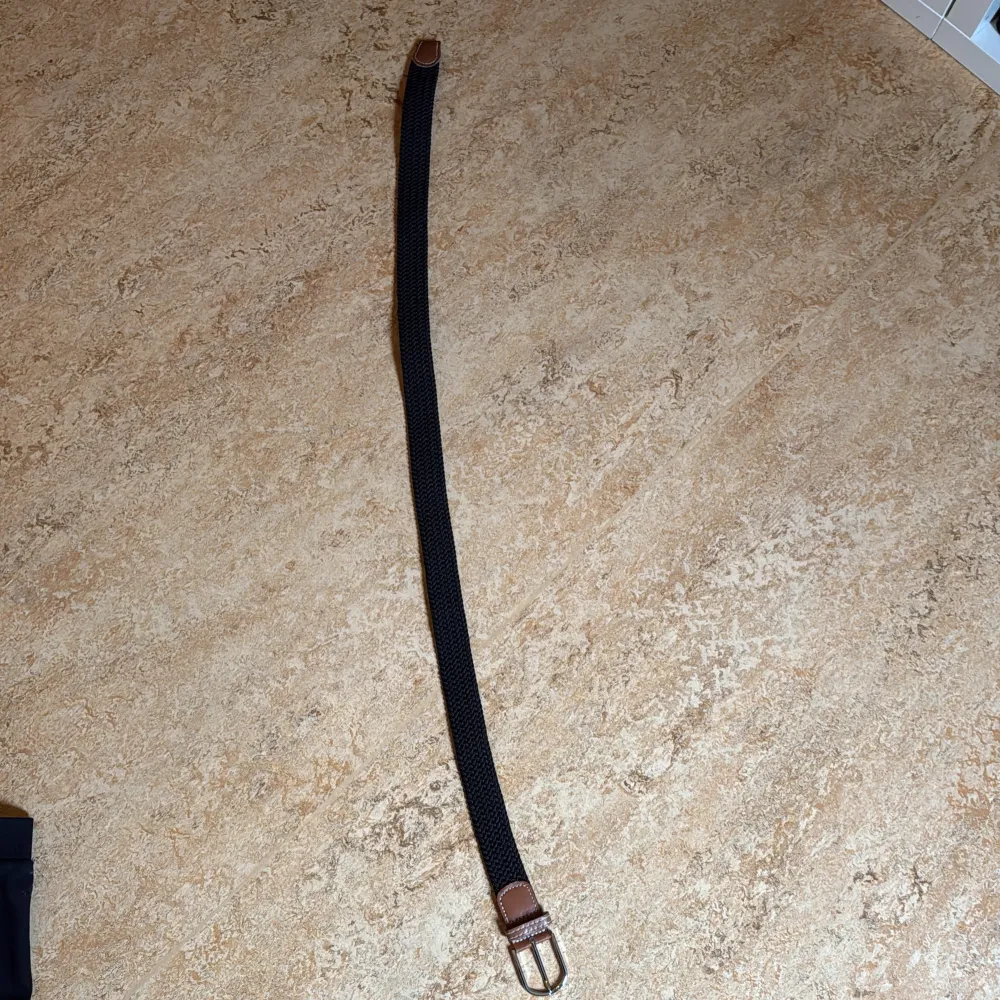 Snyggt svart flätat bälte med bruna läderdetaljer och silverfärgat spänne. Perfekt för att ge en stilren touch till din outfit. Bältet har en klassisk design med kontrasterande sömmar på läderdetaljerna.. Asusteet.