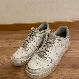 Vita Nike Air Force sneakers - Säljer ett par klassiska vita Nike Air Force sneakers. De har en ikonisk design med snörning och perforerade detaljer på ovansidan för ventilation. Perfekta för en stilren look. De är lite slitna där bak men man tänker inte på det 