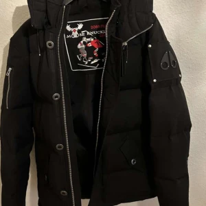 Svart dunjacka från Moose Knuckles - Snygg svart dunjacka från Moose Knuckles med dragkedja och knappar. Jackan har en cool design med en logotyp på ärmen och en quiltad look. Perfekt för kalla dagar med sin varma och stiliga stil.