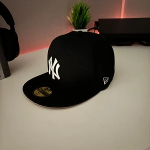 Svart keps från New Era - Snygg svart keps från New Era med broderad vit logga framtill. Klassisk 59FIFTY-modell med platt skärm och klistermärke kvar på skärmen. Perfekt för en stilren look.