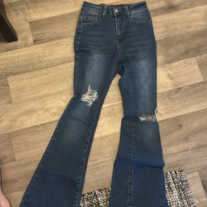 Blå bootcut jeans - Snygga blå bootcut jeans med hög midja och slitningar på låren. Perfekta för en avslappnad stil med en modern touch. Klassisk femficksdesign och knappgylf.