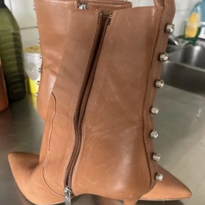 Bruna boots med dragkedja och nitar - Snygga bruna boots i skinn med dragkedja på sidan och dekorativa nitar längs kanten. De har en spetsig tå och en elegant klack, perfekt för att lyfta din stil.