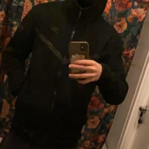 Svart Tech fleece från Nike - Säljer en svart jacka från Nike med dragkedja och huva. Jackan har en diskret logga på bröstet och är perfekt för en sportig look. Den har långa ärmar och en stilren design.