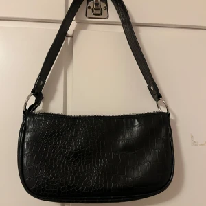 Svart axelväska från H&M Divided - Snygg svart axelväska i syntet med krokodilpräglat mönster från H&M Divided. Väskan har en praktisk dragkedja upptill och en justerbar axelrem. Perfekt för att ge din outfit en stilren touch.