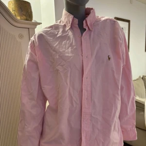 Rosa skjorta från Ralph Lauren - Säljer en klassisk rosa skjorta från Ralph Lauren. Skjortan har en broderad logotyp på bröstet och knappar framtill. Perfekt för en stilren look.