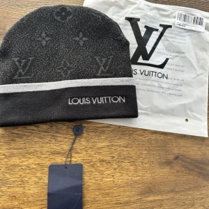 Svart mössa  - Snygg svart mössa med grått mönster och vit rand. Mössan har det ikoniska logan och är perfekt för att hålla dig varm med stil. 