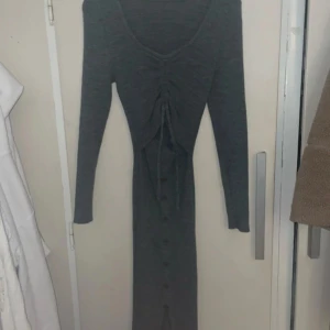 Grå långklänning med knappar - Snygg grå långklänning med knappar framtill och knytning i midjan. Klänningen har långa ärmar och är tillverkad i ett mjukt stickat material. Perfekt för en stilren look.