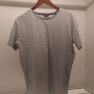 Grå t-shirt från GANT - Säljer en stilren grå t-shirt från GANT med en liten broderad logga på bröstet. T-shirten har en klassisk rund hals och korta ärmar. Perfekt för en avslappnad look.