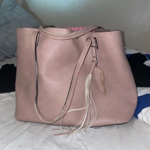 Rosa axelväska med fjäderdetalj - Snygg rosa axelväska i syntetmaterial med en cool fjäderdetalj och fransar. Väskan har en praktisk dragkedja och gott om utrymme för dina saker. Perfekt för att ge din outfit en extra touch!