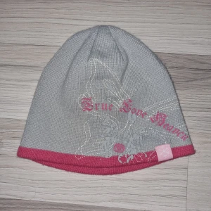 Grå och rosa mössa med text - Snygg grå mössa med rosa kant och texten 'True Love' i rosa. Mössan har ett diskret mönster och en liten etikett med en snöflinga. Perfekt för kyliga dagar.