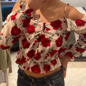 Blommig topp från H&M - Säljer en söt topp från H&M med röda rosor på vit bakrund. Toppen har långa ärmar och en volangdetalj vid midjan. Pris kan diskuteras💕