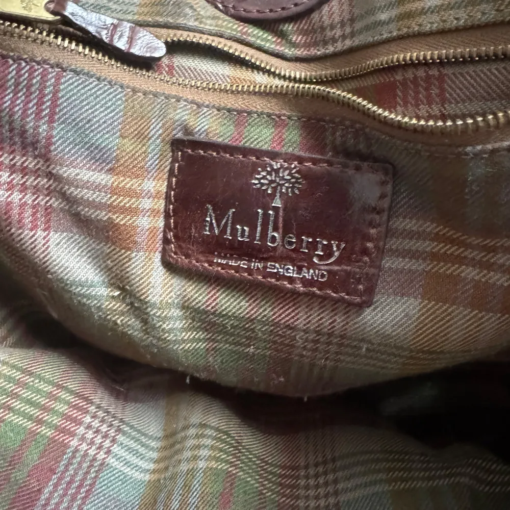 Snygg brun axelväska i skinn med krokodilpräglat mönster. Väskan har ett emblem på framsidan och en metallknapp med ett trädmotiv. Insidan är fodrad med ett rutigt tyg. Perfekt för en stilren look. ÄKTA. . Laukut & Käsilaukut.