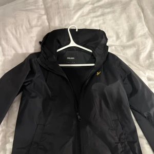 Svart vindjacka från Lyle & Scott - Snygg svart vindjacka från Lyle & Scott med dragkedja och huva. Perfekt för blåsiga dagar. Jackan har en stilren design med en liten logga på bröstet. Passar bra till både casual och sportiga outfits.