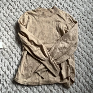 Beige stickad tröja - Säljer en mysig beige stickad tröja med långa ärmar och en hög krage. Perfekt för kyliga dagar och enkel att matcha med olika outfits. Tröjan har en avslappnad passform och ribbade detaljer vid kragen och ärmsluten.