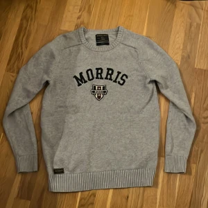 Grå stickad tröja från Morris - Säljer en stilren grå stickad tröja från Morris med svart text på bröstet. Tröjan har långa ärmar och ribbade muddar vid ärmslut och nederkant. Perfekt för en avslappnad och snygg look.