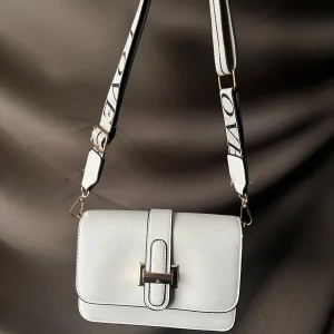 Vit/beige axelväska med textdetaljer - Snygg vit/beige axelväska med justerbar rem som har textdetaljer. Väskan har en stilren design med ett spänne framtill. Perfekt för att ge din outfit en trendig touch.