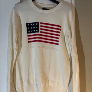 Vit gant tröja - Säljer en stilren vit stickad tröja från GANT med en amerikansk flagga och texten 'GANT NEW HAVEN' på framsidan. Tröjan har långa ärmar och en rund halsringning. Perfekt för en avslappnad och trendig look. Lappen sitter löst men går säkert att fixa, storlek M