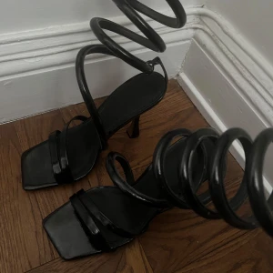 Svarta sandaler med spiralremmar från ASOS - Snygga svarta sandaler från ASOS med unika spiralformade remmar som slingrar sig upp längs benen. De har en fyrkantig tå och en elegant klack, vilket ger en modern och stilren look.