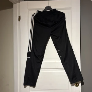 Svarta träningsbyxor med vita ränder - Snygga svarta träningsbyxor med klassiska vita ränder längs sidorna. Byxorna har en bekväm passform och elastisk midja, perfekt för sport och träning. De har även vita detaljer runt knäna för en extra touch. Det är 3 likadana byxor i samma paket här och samma storlekar på alla 3 byxorna.