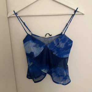 Blå transparent linnetopp från Pull&Bear - Snygg blå transparent linnetopp från Pull&Bear med justerbara axelband. Toppen har ett abstrakt mönster i olika blå nyanser och en lätt croppad passform. Perfekt för varma dagar eller som ett lager under en jacka.