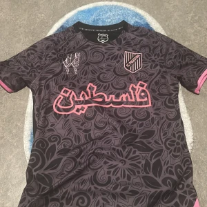 Svart och rosa fotbollströja med blommönster - Snygg fotbollströja i svart med rosa detaljer och ett unikt blommönster. Tröjan har korta ärmar och en stilren design med text och emblem på framsidan. Perfekt för dig som vill sticka ut på planen!