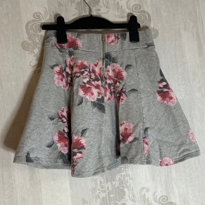 Sötaste blommiga kjolen - Säljer en söt grå kjol med rosa blommönster från H&M Divided. Kjolen har en kort längd och är perfekt för vår och sommar. Dragkedja bak. Sparsamt använd 
