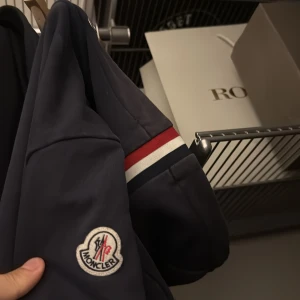 Mörkblå zip hoodie från Moncler - Säljer en snygg mörkblå zip hoodie från Moncler med klassisk logga på ärmen. Zip hoodien har en dragkedja framtill och detaljer i rött och vitt. Perfekt för en stilren look. Kom privat för mer bilder! OBS säljer inte mindre än 1000, den är äkta+ kvitto ingår