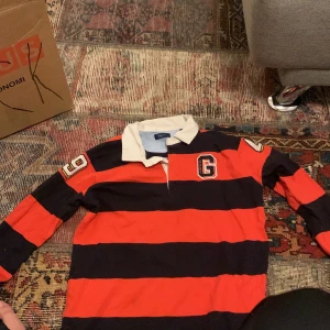 Randig pikétröja från Gant - Säljer en cool randig pikétröja från Gant i rött och svart med vita detaljer. Tröjan har långa ärmar och en klassisk krage. Siffrorna 49 och 19 pryder ärmarna och ryggen har ett stort Gant-tryck. Perfekt för en sportig look!