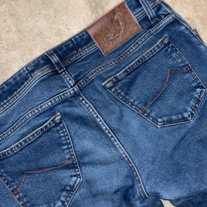 Blå jeans från Jacob Cohen - Snygga blå jeans från Jacob Cohen med klassisk femficksdesign och knappgylf. De har en broderad detalj på insidan av gylfen och en läderpatch med logga på baksidan. Perfekta för en stilren look. Och har ett litet hål nedanför gylfen.  Pris kan diskuteras!