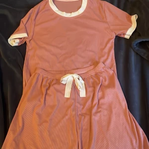 Rosa ribbade shorts med matchande t-shirt - Säljer ett set med rosa ribbade shorts och matchande t-shirt. Shortsen har en bekväm passform med vit knytning i midjan och vita detaljer vid bensluten. T-shirten har vita kanter vid ärmar och hals. Perfekt för en avslappnad dag.