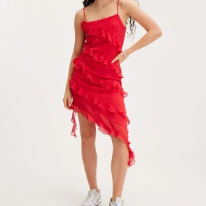 Röd klänning med volanger - Snygg röd klänning med volanger och smala axelband. Klänningen har en asymmetrisk design med en dekorativ blomma på axeln. Bara använd vid ett tillfälle. I mycket bra skick!🌹❤️