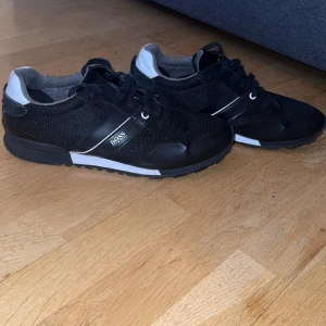 Svarta sneakers från Boss - Snygga svarta sneakers från Boss med vita detaljer. Skorna har snörning och en stilren design som passar perfekt för en casual look. De är tillverkade i en kombination av syntet och skinn för en hållbar och bekväm känsla.