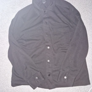 Svart overshirt med knappar - Säljer en stilren svart overshirt med knappar framtill och två stora bröstfickor. Perfekt för lager-på-lager-stil och passar till många olika outfits. Enkelt och snyggt plagg för både vardag och mer uppklädda tillfällen.