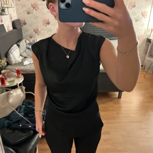 Svart ärmlös topp - Snygg svart ärmlös topp med en elegant och enkel design. Perfekt för en stilren look.Glansigt material och dragkedja i ryggen.