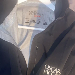 Oscar Jacobson jacka/windbreaker - Säljer en stilren svart jacka från Oscar Jacobson i storlek S. Jackan har dragkedja framtill och två praktiska fickor med dragkedjor. Perfekt för höst och vår med sin snygga design och funktionella detaljer.