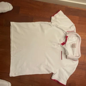 Vit kortärmad t-shirt från Guess - Säljer en vit kortärmad t-shirt från Guess med röda detaljer vid krage och ärmslut. T-shirten har en slim fit passform och är designad i Kalifornien. Perfekt för en stilren och avslappnad look.