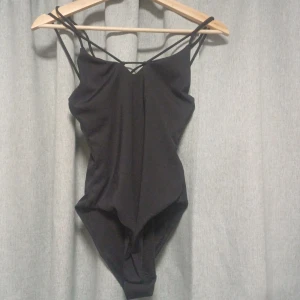Svart bodysuit - Svart body med smala band och en kors liknande design på baksidan. Försluten gren. Helt oanvänd men har ingen prislapp