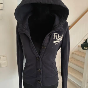 Mörkblå huvtröja från Abercrombie & Fitch - Säljer en snygg mörkblå huvtröja från Abercrombie & Fitch med broderad logga på bröstet. Tröjan har knappar framtill och en mysig huva. Perfekt för en avslappnad stil.