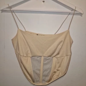 Beige korsettopp med meshdetaljer - Snygg beige korsettopp med tunna axelband och meshdetaljer framtill. Perfekt för en stilren och modern look. Garderobsrensning så kika gärna på övriga auktioner.