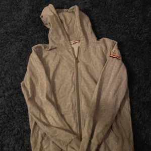 Grå huvtröja från Moncler - Säljer en stilren grå huvtröja från Moncler med dragkedja och broderad logga på ärmen. Tröjan har en klassisk design med ribbade detaljer och en bekväm passform. Den är ganska gammal och har fått den av en vän så äkthet kan inte garanterats därför priset, snörrerna har också trillat av, det är storlek xxl i barn storlek så tröjan passar mig som vanligt vis har m/L skriv för frågor eller annat privat